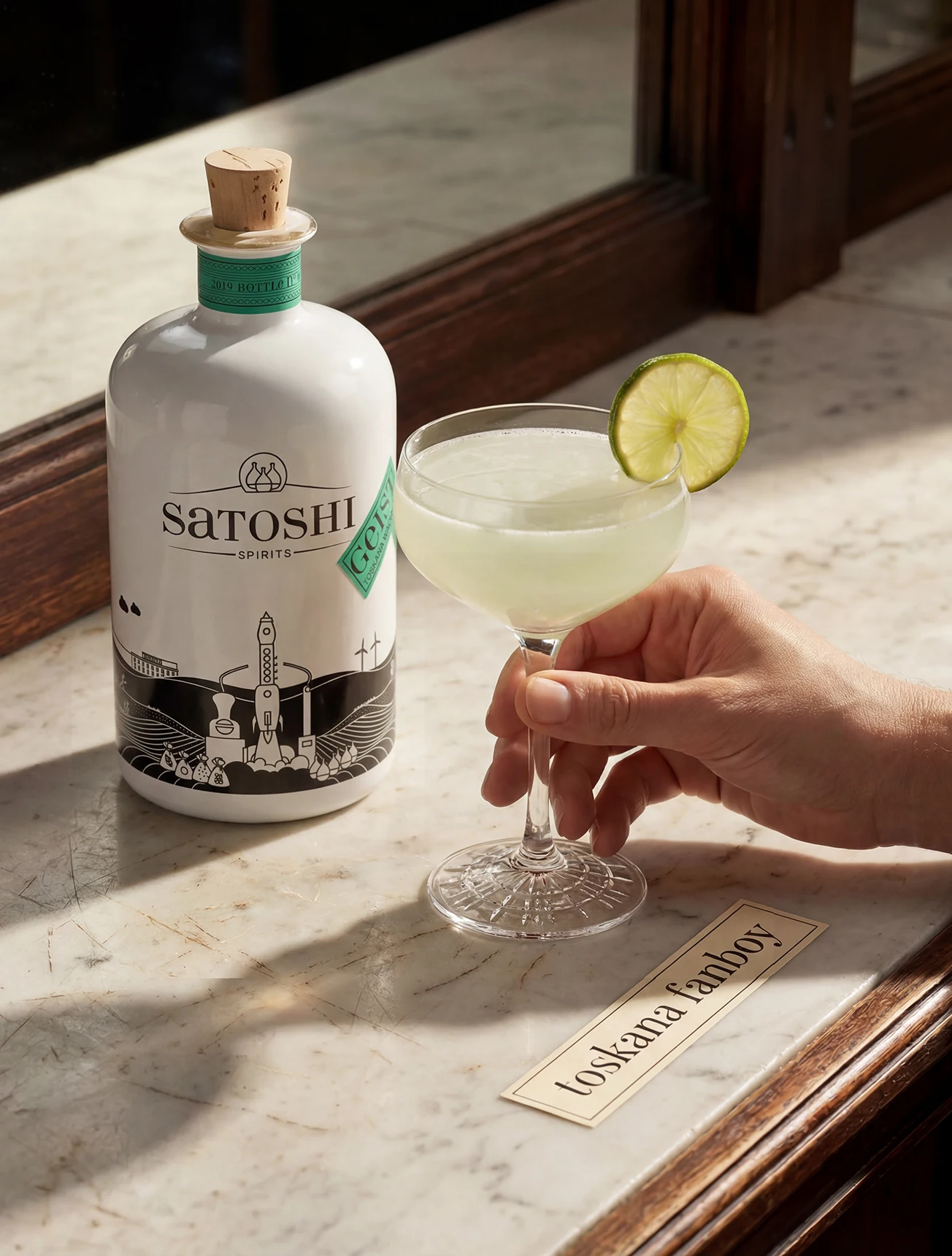 Männlich gelesene Hand greift nach Gimlet Cocktail neben der Satoshi Flasche des Geist vom toskanischen Wacholder